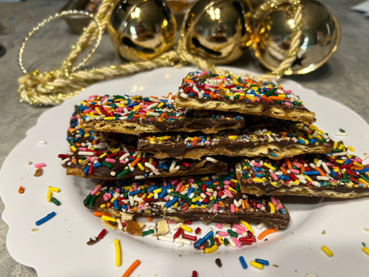 A white  plate of Christmas Maple crack with jingle bells in the background. The Maple Crack is covered in chocolate and has multi colored sprinkles. 